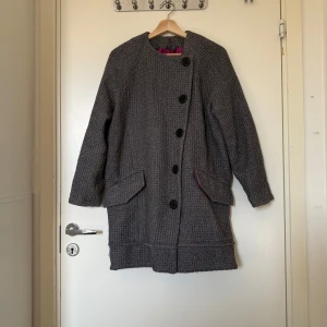 Grå kappa från Odd Molly i ullmix - Säljer en grå kort kappa från Odd Molly med snyggt vävt mönster och stora svarta knappar. Fodret är i en cool cerise färg som poppar till looken. Kappan har två fickor framtill och är långärmad, perfekt för höst och vinter. Storlek 0 som enligt deras storleksguide motsvarar en storlek XS, men den har bröstmåttet 50cm. Så den är rymlig i storlek. Kanske S/M. Fint skcik, men den kan vara lite stickig på utsidan, materialet är 50% ull, 50% viskos. 
