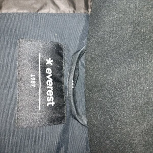 Svart parkasjacka från Everest - Säljer en svart parkasjacka från Everest med huva och fuskpäls. Jackan har stora fickor framtill, dragkedja och knappar. Perfekt för kalla dagar med sin långa modell och varma foder. Klassisk och stilren design som funkar till det mesta.