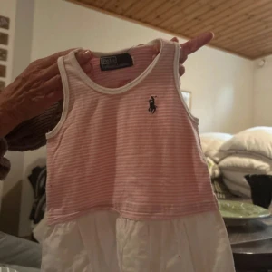 Barn klänning Polo Ralph Lauren - Supersöt klänning från Polo Ralph Lauren med rosa och vita ränder upptill och vit kjoldel. Ärmlös modell med klassisk logga broderad på bröstet. Perfekt för varma dagar och har en lätt och luftig känsla.