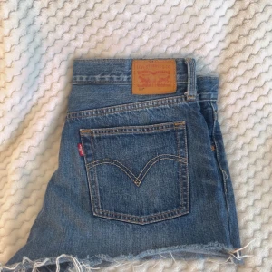 Levi's blå jeansshorts med fransig kant - Snygga blå jeansshorts från Levi's med klassisk femficksdesign och fransig rå kant nedtill. De har hög midja, Levi's patch bak och den ikoniska röda etiketten på fickan. Perfekta för sommarens alla äventyr och har en loose passform.