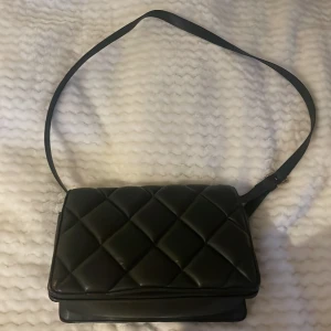 Svart quiltad axelväska i skinn - Snygg svart axelväska med quiltad design och bred axelrem. Väskan har ett stilrent rutmönster och är tillverkad i skinn. Perfekt storlek för att få plats med allt du behöver och har ett fack på insidan för småsaker. Enkel att matcha med olika outfits.