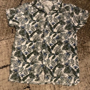 Vit blommig skjorta från D.M.D , 158/164 - Snygg kortärmad skjorta från D.M.D med tropiskt blommigt mönster i grönt och blått på vit botten. Skjortan har klassisk krage och vita knappar framtill. Perfekt för dig som gillar färg och mönster!