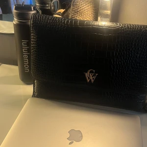Svart datafodral  - Säljer en svart kuvertväska från CW med croco-mönstrad yta i skinnimitation och guldfärgad logga framtill. Väskan har magnetlås och ett rymligt foder med CW-mönster. Perfekt för att bära laptop eller viktiga papper med stil.