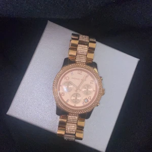 Guldig Michael Kors klocka med stenar - Snygg klocka från Michael Kors i guld med glittriga stenar på både boett och armband. Urtavlan är ljusrosa med tre mindre visartavlor och datumvisning. Länken är bred och har detaljer med små stenar för extra bling.