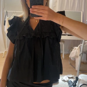 Svart blus med volangdetaljer - Supersöt svart blus med v-ringning och volangdetaljer över axlar och bröst. Luftig passform och korta ärmar som ger en avslappnad men ändå chic vibe. Perfekt att styla med jeans eller kjol för en trendig look.
