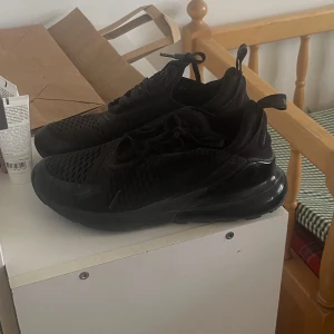 Svarta Nike Air Max 270 sneakers - Säljer ett par svarta Nike Air Max 270 sneakers med snygg mesh-överdel och synlig Air-enhet i hälen. Skorna har snörning och en sportig, modern look som passar perfekt till streetwear. Swoosh-loggan på sidan och platt sula för skön komfort. Passar mig som har storlek 39/40 och breda fötter då de är herrmodell men passar lika väl på dam. 