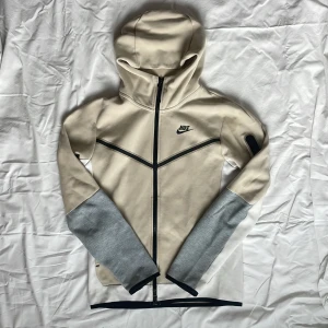 Nike beige hoodie med dragkedja - Nike hoodie i beige med svarta detaljer och hel dragkedja. Ärmarna har grå muddar och en liten ficka med dragkedja på ena ärmen. Tröjan har huva och svart Nike-logga på bröstet. Perfekt för chill eller träning.