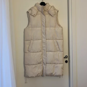 Vit/Beige lång dunväst från Violet Romance - Lång, quiltad vit/beige dunväst från Violet Romance med huva och tryckknappar framtill. Västen har två snedställda fickor och dragsko nertill för justerbar passform. Perfekt för lager-på-lager och en clean look.