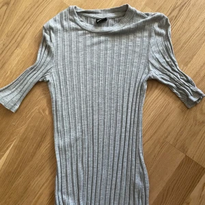 Grå ribbad kortklänning Gina Tricot - Supersnygg grå ribbad klänning från Gina Tricot med korta ärmar och rund hals. Klänningen är figurnära och har vertikala ribbade ränder som ger en cool struktur. Perfekt för dig som gillar enkel men trendig stil.