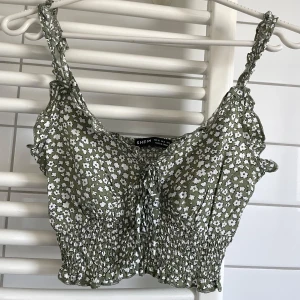 Grön blommig croppad topp  - Supersöt grön croppad topp med vitt blommönster, smockad midja och volangdetaljer. Axelbandsmodell med knyt framtill och rynkade axelband. 