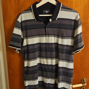 Randig pikétröja från Dressmann XL - Snygg pikétröja från Dressmann i storlek XL med breda horisontella ränder i marinblått, grått och vitt. Klassisk krage med vit kant, korta ärmar och bröstficka. Perfekt för en chill och stilren look.