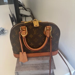 Louis Vuitton Monogram Alma BB handväska - Säljer en ikonisk Louis Vuitton Alma BB handväska i klassiskt monogrammönster. Väskan är brun med ljusbruna skinnhandtag och detaljer i guldmetall. Den har dragkedja med lås och en avtagbar axelrem. Köpt i Newyork 2023 kvitto orginalförpackning medföljer kortat in den 2 cm hos skräddare se bild inget som syns nypris 21.000 sek inkl. Skattavgift  