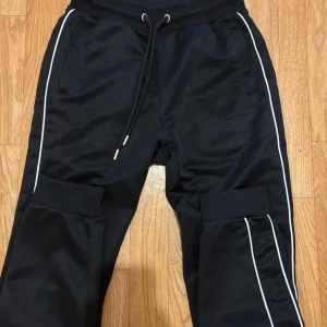 Svarta mjukisbyxor Supply & Demand - Svarta mjukisbyxor från Supply & Demand med vita ränder längs benen och snörning i midjan. Byxorna har ribbade muddar vid bensluten och praktiska fickor. Perfekta för en chill och sportig look.