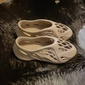 Beige Adidas Yeezy Foam Runner - Säljer ett par beige Adidas Yeezy Foam Runner med futuristisk design och många öppna hål för extra ventilation. Skorna är tillverkade i syntetmaterial och har en platt sula. Perfekt för dig som gillar unik stil och komfort.