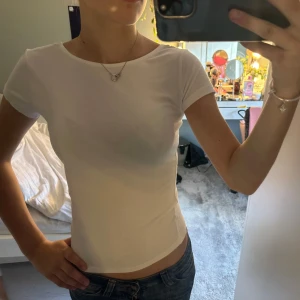 Vit t-shirt med rosett och djup rygg - Supersöt vit t-shirt med korta ärmar och en djup, öppen rygg som pryds av en stor, glansig rosett i satin. T-shirten är figurnära och gjord i mjuk bomull, perfekt för dig som gillar enkla men ändå unika detaljer.