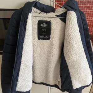 Mörkblå pufferjacka från Hollister S - Säljer en mörkblå pufferjacka från Hollister med vit teddyfoder på insidan. Jackan har huva, dragkedja framtill och diskret logga på bröstet. Perfekt för kalla dagar och riktigt skön att bära. Snygg och enkel design som funkar till det mesta.