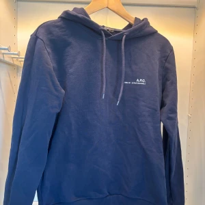 Marinblå hoodie från A.P.C. - Säljer en stilren marinblå hoodie från A.P.C. med vit logga på bröstet. Tröjan har huva med snörning och är tillverkad i mjuk bomull. Perfekt för dig som gillar minimalistisk och clean streetstyle. Ny pris cirka 2700.
