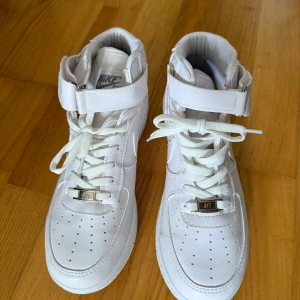 Nike Air Force 1 Mid vita sneakers - Nike Air Force 1 Mid sneakers i helvitt skinn med klassisk swoosh på sidan och broderad logga på hälen. Skorna har snörning, kardborreband vid ankeln och perforerad tå för extra ventilation. Platt sula med tydlig Air-dämpning och rund tå. Perfekt för dig som gillar clean och tidlös streetstil.