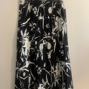 Svart och vit blommig kjol från Isolde - Snygg plisserad svart kjol med vitt blommönster från Isolde. Kjolen har en plisserad design som ger en fin struktur och rörelse. Perfekt för att ge en elegant touch till din outfit.