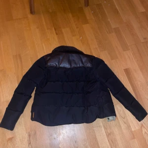 Svart dunjacka från Penfield - Säljer en svart dunjacka från Penfield med bruna läderdetaljer på axlarna. Jackan har en klassisk design med knappar framtill och en mjuk krage. Perfekt för kyliga dagar.