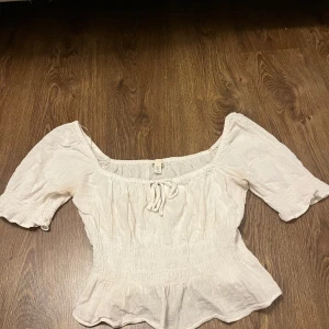 Vit blus från H&M - Säljer en söt vit blus från H&M med korta puffärmar och en smockad midja. Blusen har en knytning framtill och en lätt volang vid nederkanten. Perfekt för en somrig look! 🌼
