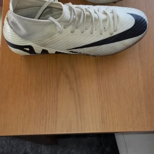 Vita fotbollsskor från Nike - Säljer ett par vita fotbollsskor från Nike med svart swoosh. Skorna har snörning och är designade för optimal prestanda på planen. Perfekta för fotbollsspelare som vill ha stil och funktionalitet.