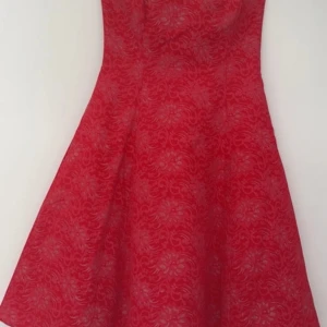 Röd cocktail klänning med korsett - Elegant röd cocktail/bal klänning med silvriga detaljer och snörning i ryggen. Knälång med dragkedja i sidan. Spets och tyll i underkjol för volym. Storlek M men går att storleksjustera tack vare snörningen i ryggen.