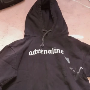 Svart hoodie med texten 'adrenaline' - Säljer en svart hoodie med texten 'adrenaline' i vitt på bröstet. Den har en klassisk design med huva och dragsko. Perfekt för en avslappnad stil. För både tjejer och killar