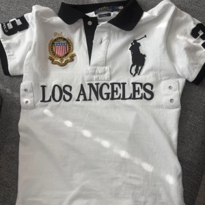 Chief keef Ralph Lauren - Snygg vit pikétröja från Ralph Lauren med svart krage och broderad logga. Stort 'Los Angeles'-tryck på framsidan och siffran 3 på ärmarna och ryggen. Perfekt för en sportig look!