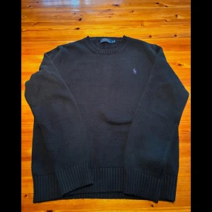 Svart tröja från Polo Ralph Lauren - Säljer en klassisk svart tröja från Polo Ralph Lauren i mycket bra skick med lila broderad logga på bröstet. Tröjan har en rund halsringning och långa ärmar, perfekt för en stilren look. 