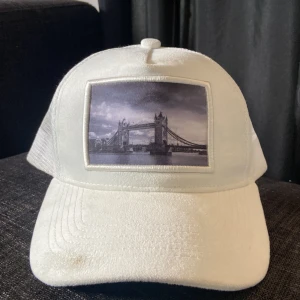 Vit keps med Tower Bridge-tryck - Snygg vit keps från Gårda med ett svartvitt tryck av Tower Bridge på framsidan. Kepsen har ett nätmaterial på baksidan för extra ventilation och en justerbar rem för perfekt passform.