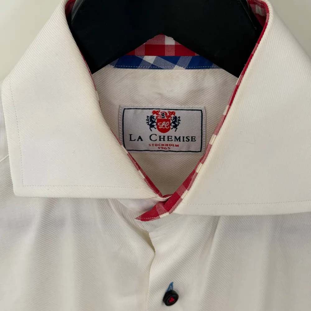 Snygg vit skjorta från La Chemise med unika detaljer. Den har en rutig insida i rött och vitt samt svarta knappar. Perfekt för en stilren look med en twist.. Paidat.