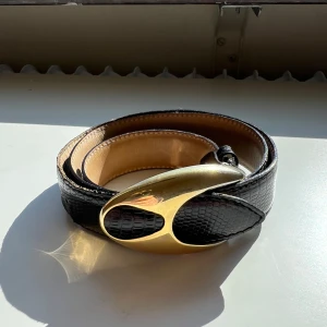 Vintage Bälte Svart - Elegant svart vintage bälte i skinn med ett unikt guldspänne. Tillverkat i Italien, detta bälte har en lyxig känsla och passar perfekt för att lyfta vilken outfit som helst. 