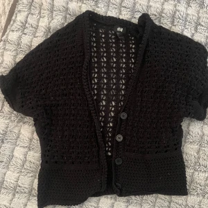 Svart stickad kofta från H&M - Säljer en svart stickad kofta från H&M med kortärmad design och knappar framtill. Perfekt för lager-på-lager och ger en stilren look. Passar bra till både vardag och mer uppklädda tillfällen.