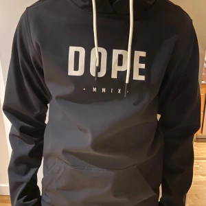 Svart jacka från Dope - Snygg svart jacka från Dope med vit text på framsidan. Den har en justerbar huva med vita snören och en praktisk magficka. Perfekt för en avslappnad stil i skidbacken! Hör av er vid intresse eller frågor! 
