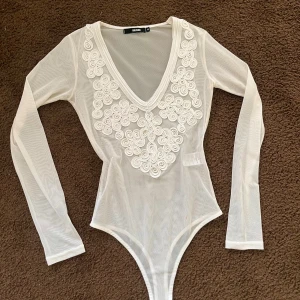 Vit långärmad bodysuit från bikbok - Säljer en superfin vit bodysuit från Bikbok med v-ringning och dekorativa spetsdetaljer framtill. Den är långärmad och tillverkad i ett transparent material som ger en stilren look. Perfekt för att styla med jeans eller kjol.