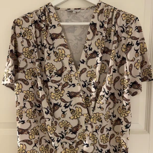 Mönstrad blus med v-ringning - Kortärmad blus med ett livfullt mönster i beige, gul och svart. Wrap-modell med v-ringning. Passar utmärkt för både vardag och speciella tillfällen. Från MQ.
