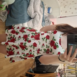 Vit blommig klänning från H&M Divided - Säljer en söt vit klänning med röda rosor från H&M Divided. Klänningen har en kort längd och är ärmlös, perfekt för sommarens varma dagar. Den har en skön passform och ett härligt blommigt mönster som verkligen sticker ut.