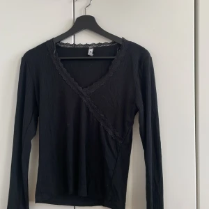 Svart långärmad topp med spetsdetaljer - Snygg svart långärmad topp med spetsdetaljer vid halsringningen. Perfekt för en stilren look. Toppen har en omlott-design framtill som ger en elegant touch. 