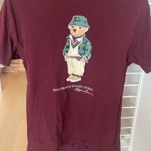 Vinröd t-shirt med Polo Bear från Ralph Lauren - Säljer en vinröd t-shirt från Ralph Lauren med en cool Polo Bear-tryck på framsidan. T-shirten är kortärmad och gjord i mjuk bomull. Perfekt för en avslappnad stil!
