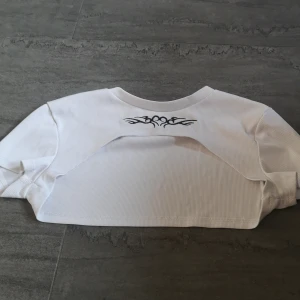 Vit ribbad bolerotopp från FB Sister - Säljer en vit ribbad bolerotopp från FB Sister med ett coolt svart tribal-mönster på framsidan. Toppen är kortärmad och har en stilren design som passar perfekt för en trendig look. Perfekt för att lägga till en edgy touch till din outfit! Man kan ha linne eller andra tröjer under, inte rekommenderad att vara gå i den utan något under!!😂❤️