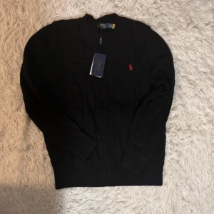 Svart stickad tröja från Polo Ralph Lauren - Säljer en stilren svart stickad tröja från Polo Ralph Lauren med det klassiska röda logotypen broderad på bröstet. Tröjan har en ribbad rund hals och långa ärmar, perfekt för kyligare dagar. Helt ny med lapp, o qr kod!