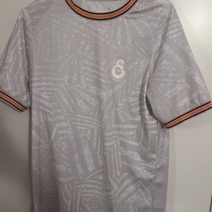 Galatasaray fotbollströja - Snygg grå fotbollströja med subtilt mönster och korta ärmar. Tröjan har en rund halsringning med ränder i orange och svart. Perfekt för sportiga tillfällen eller som en del av en avslappnad stil.