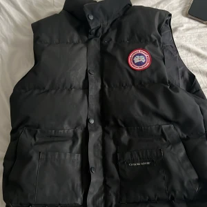 ÄKTA Svart dunväst från Canada Goose - Säljer en svart dunväst från Canada Goose med deras ikoniska märke på bröstet. Västen har knappar framtill och praktiska fickor. Perfekt för kyliga dagar.