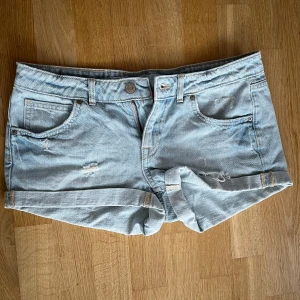 Ljusblå jeansshorts - Säljer dessa ljusblå lågmidjade jeansshorts med slitna detaljer. Shortsen har fem fickor och en knappgylf. Jag säljer då de inte var min storlek. Jag skulle säga att de passar storlek S.
