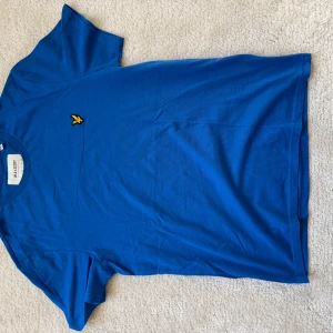 Blå t-shirt från Lyle & Scott - Säljer en snygg blå t-shirt från Lyle & Scott i ekologisk bomull. Den har en liten broderad logga i gult på bröstet och är kortärmad. Perfekt för en avslappnad stil. Storlek M.