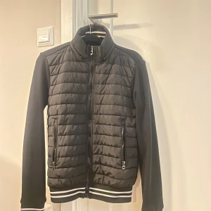 Svart quiltad jacka med dragkedja - Snygg svart jacka med quiltad framsida och dragkedja. Den har långa ärmar och ribbade muddar med vita ränder vid ärmslut och nederkant. Perfekt för en stilren look.