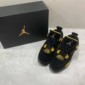 Svarta och gula Jordan 4 - Snygga Nike Air Jordans i svart och gult med ikonisk design. Skorna har en robust konstruktion med snörning och en bekväm passform. Perfekta för dig som vill ha en stilren look med en touch av färg.