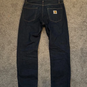 Mörkblå jeans från Carhartt - Säljer ett par mörkblå jeans från Carhartt med klassisk femficksdesign och logotyp på bakfickan. Jeansen har en rak passform och är tillverkade i slitstarkt denim. Perfekta för en avslappnad stil.