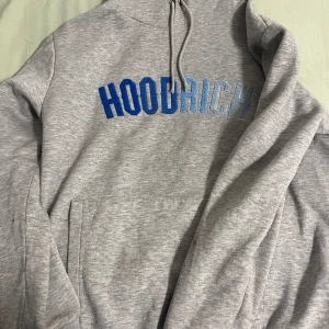 Grå hoodie från Hoodrich - Säljer en grå hoodie från Hoodrich med blå text på bröstet. Tröjan har en stor ficka framtill. Perfekt för en avslappnad stil.